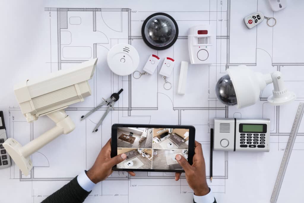 Installation Système Sécurité : Plan, Caméras et Alarme Mains tenant une tablette affichant quatre flux de caméras de sécurité, posée sur un plan d'architecte avec divers équipements d'alarme.